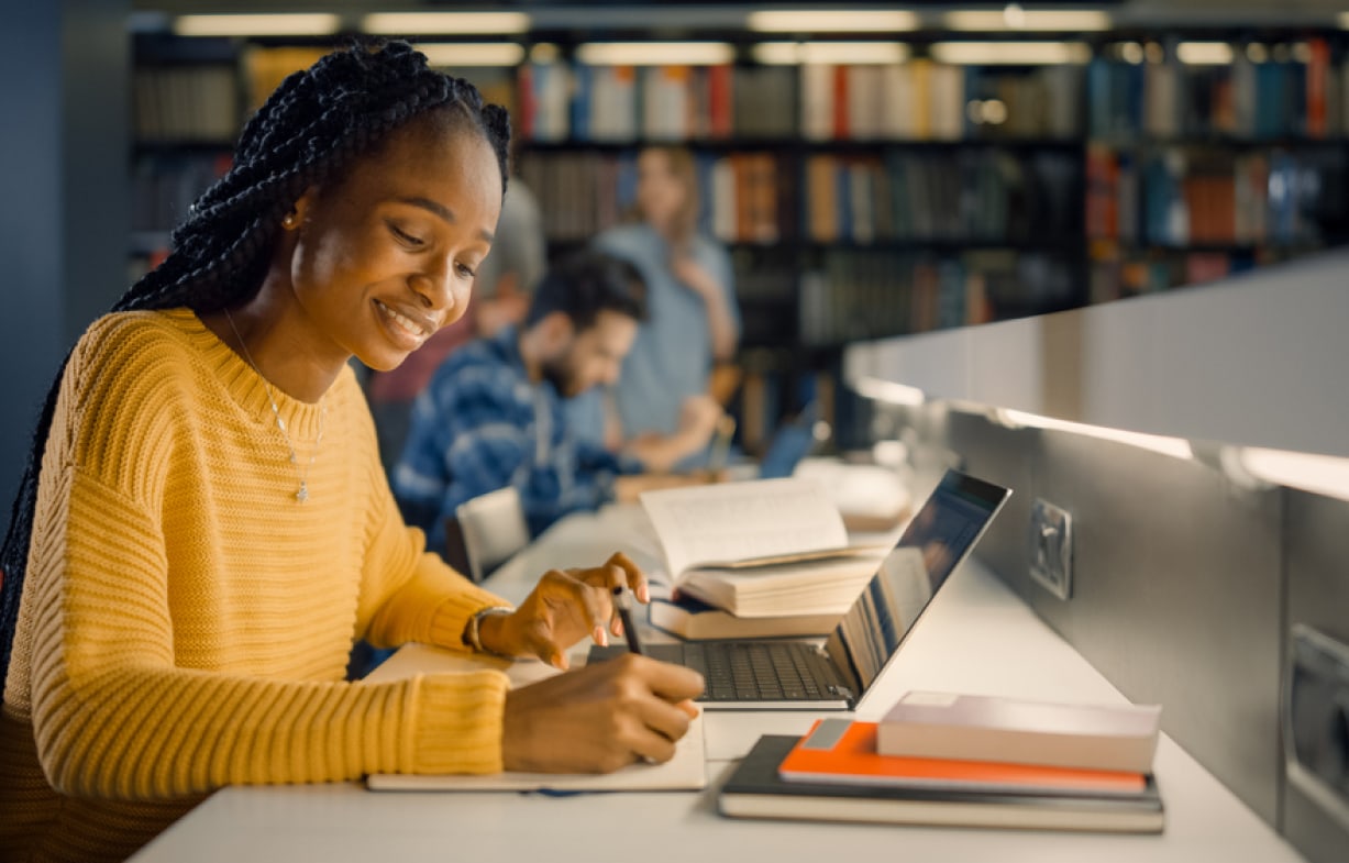 A young woman sitting at a table in a library, focused on her laptop.