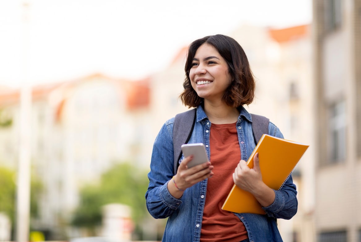 A young woman smiles while holding a folder and glancing at her phone, multitasking with ease.